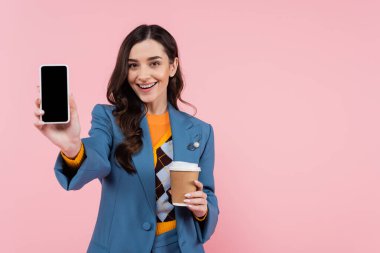cheerful young woman in blue blazer holding smartphone with blank screen and paper cup isolated on pink 