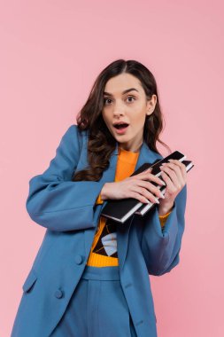 shocked and brunette manager in blue blazer holding notebooks isolated on pink 