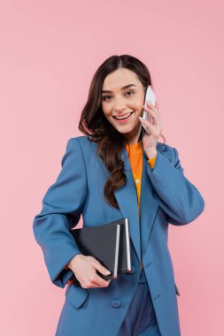 brunette manager in blue blazer holding notebooks and talking on smartphone isolated on pink 