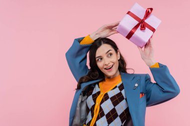 cheerful young woman in blue blazer holding wrapped present above head isolated on pink 