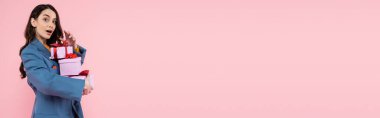 astonished woman in blue blazer holding gift boxes and looking at camera isolated on pink, banner