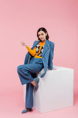 full length of trendy brunette woman in blue suit posing near white cube and looking at camera on pink background