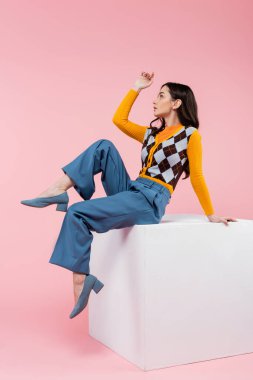 side view of young woman in stylish cardigan and blue pants looking away while sitting on white cube on pink background