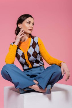 full length of woman in blue pants and orange cardigan with argyle pattern sitting with crossed legs on white cube isolated on pink