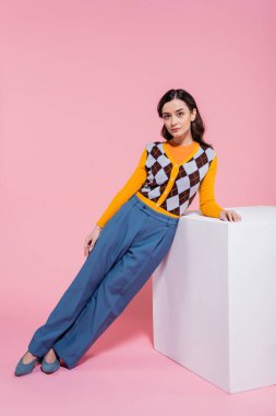 full length of trendy woman in blue trousers and orange cardigan with argyle pattern leaning on white cube on pink background