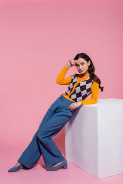 full length of pretty woman in trendy cardigan and blue pants leaning on white cube on pink background