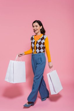 full length of impressed woman in trendy cardigan and blue pants walking with shopping bags on pink background clipart