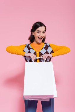 excited woman in orange cardigan with argyle pattern holding white shopping bags and looking at camera on pink background