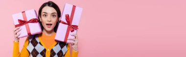 surprised woman in trendy cardigan holding presents and looking at camera isolated on pink, banner