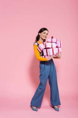 full length of cheerful woman in blue pants holding plenty of presents and looking at camera on pink background