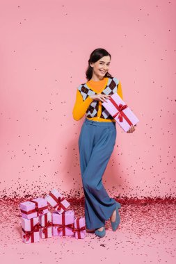 full length of pleased woman posing with present near festive confetti and gift boxes on pink background