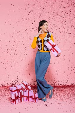 full length of surprised woman in trendy outfit standing with present near gift boxes and festive confetti on pink background