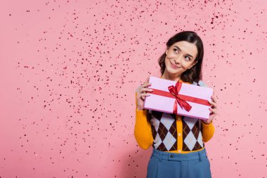 happy and dreamy woman in stylish clothes holding present and looking away near festive confetti on pink background