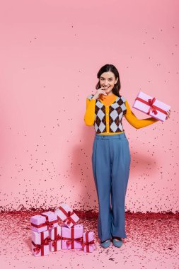 pleased woman in blue pants holding present and looking at camera near confetti and gift boxes on pink background