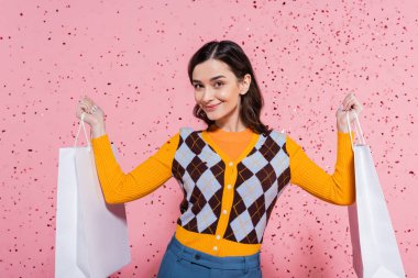 pleased woman in stylish cardigan holding shopping bags and smiling at camera on pink background