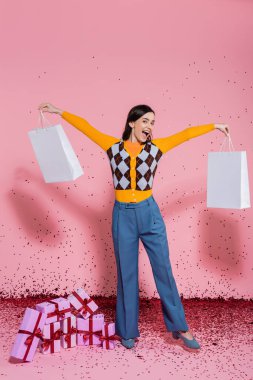 excited woman in stylish clothes posing with white shopping bags near festive confetti and gift boxes on pink background