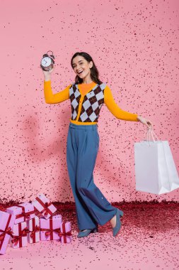 joyful and fashionable woman posing with alarm clock and shopping bags near confetti and gift boxes on pink background