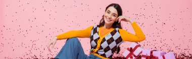 cheerful woman in fashionable attire posing near gift boxes and festive confetti on pink background, banner