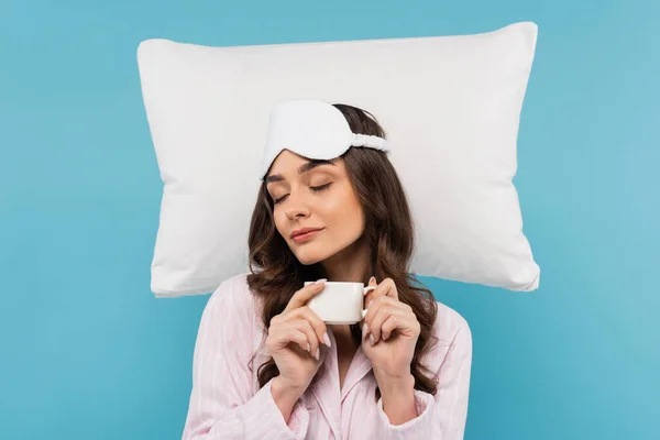 stock image sleepy woman in pajamas and night mask holding cup of coffee near white flying pillow isolated on blue