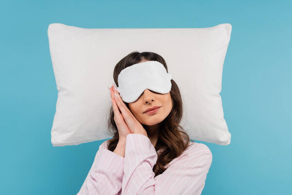молодая женщина в пижаме и ночной маске отдыхает на белой подушке, изолированной на голубой 