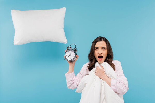 шокированная молодая женщина в пижаме держа одеяло и винтажные будильники возле летающей подушки на голубой 