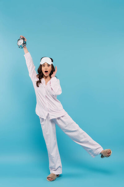 full length of shocked young woman in night mask and sleepwear holding alarm clock on blue 