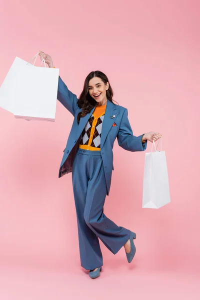 stock image full length of positive young woman in blue pantsuit holding shopping bags on pink 