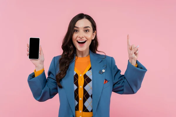 Stock image excited young woman in blue blazer holding smartphone with blank screen and pointing up isolated on pink 