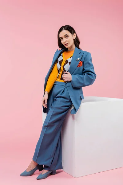 full length of brunette woman in blue suit posing near white cube and looking at camera on pink background