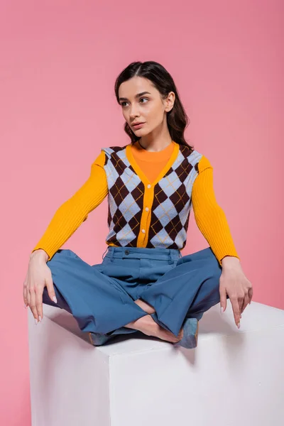 full length of brunette model in blue pants sitting with crossed legs on white cube on pink background