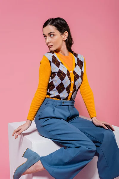 brunette woman in blue pants and orange cardigan with argyle pattern sitting on white cube and looking away isolated on pink