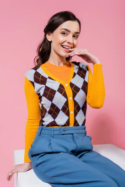coquettish woman in blue pants and orange cardigan with argyle pattern sitting on white cube on pink background