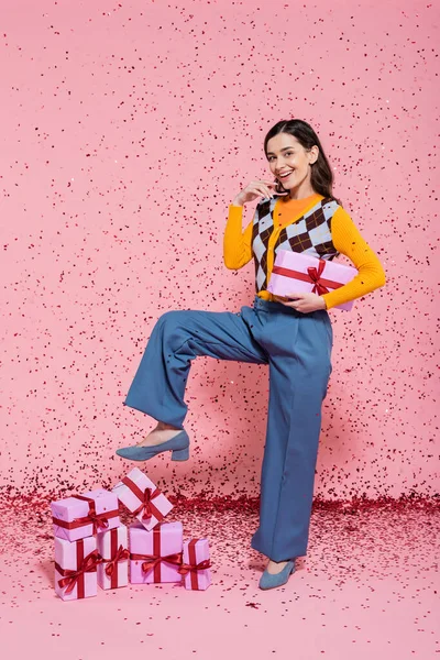 joyful woman in blue pants holding gift box while posing near pile of presents and confetti on pink background