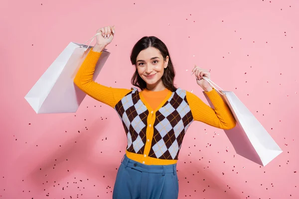 stock image joyful woman in orange cardigan with argyle pattern posing with white shopping bags on pink background