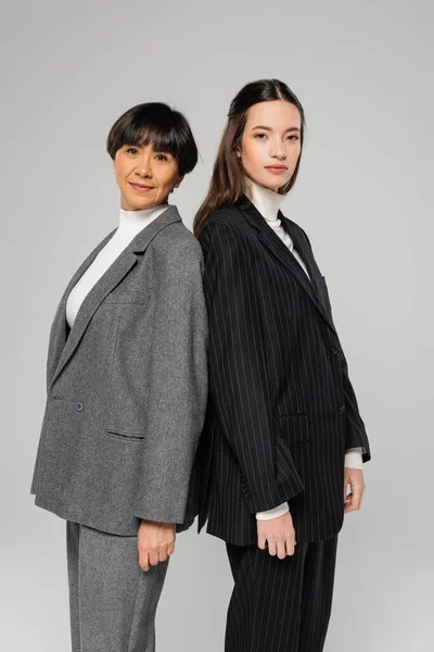 stock image asian mother and daughter in stylish suits looking at camera while standing back to back isolated on grey