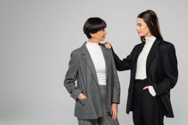 brunette woman in black suit touching shoulder of elegant asian mother isolated on grey