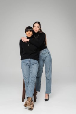 full length of fashionable woman embracing middle aged asian mother while posing on grey background