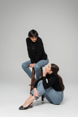 full length of brunette woman looking at stylish asian mother sitting on wooden stool on grey background