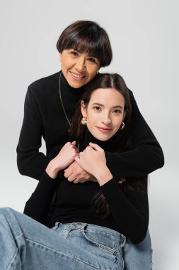 joyful asian woman hugging young and stylish daughter while looking at camera on grey background