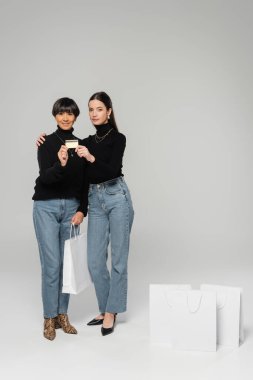 full length of asian mother and daughter in stylish clothes showing credit card near white shopping bags on grey background