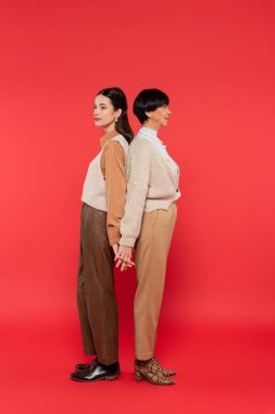 full length of asian mother and daughter in stylish casual attire holding hands while standing back to back on coral background