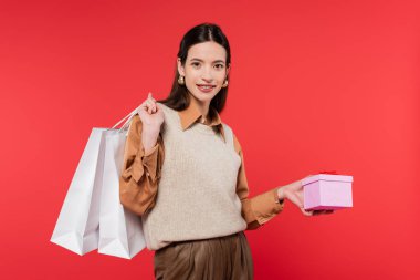young woman in stylish casual attire holding present and shopping bags while smiling at camera isolated on coral