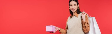 cheerful and stylish asian woman holding gift box and shopping bags isolated on coral, banner