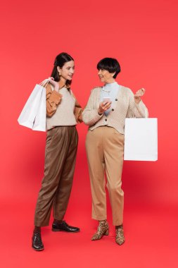 full length of trendy asian mother and daughter with mobile phone and shopping bags looking at each other on coral background