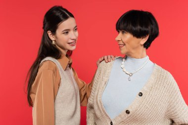 cheerful asian mother and daughter looking at each other isolated on coral 