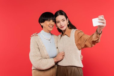 young asian woman in knitted vest taking selfie with happy mother isolated on coral 