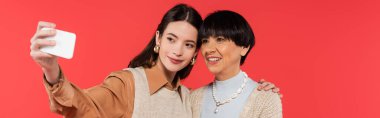 young asian woman in knitted vest taking selfie with happy mother isolated on coral, banner 