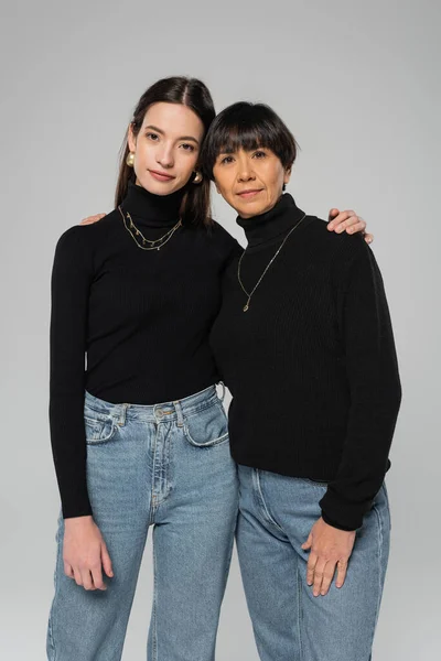 asian mother and daughter in black turtlenecks and blue jeans looking at camera while standing isolated on grey