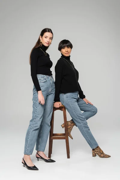 full length of asian mother and daughter in blue jeans and black turtlenecks posing near wooden stool on grey background
