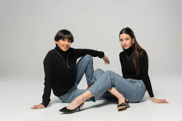 full length of asian mother and daughter in blue jeans and black turtlenecks sitting and looking at camera on grey background